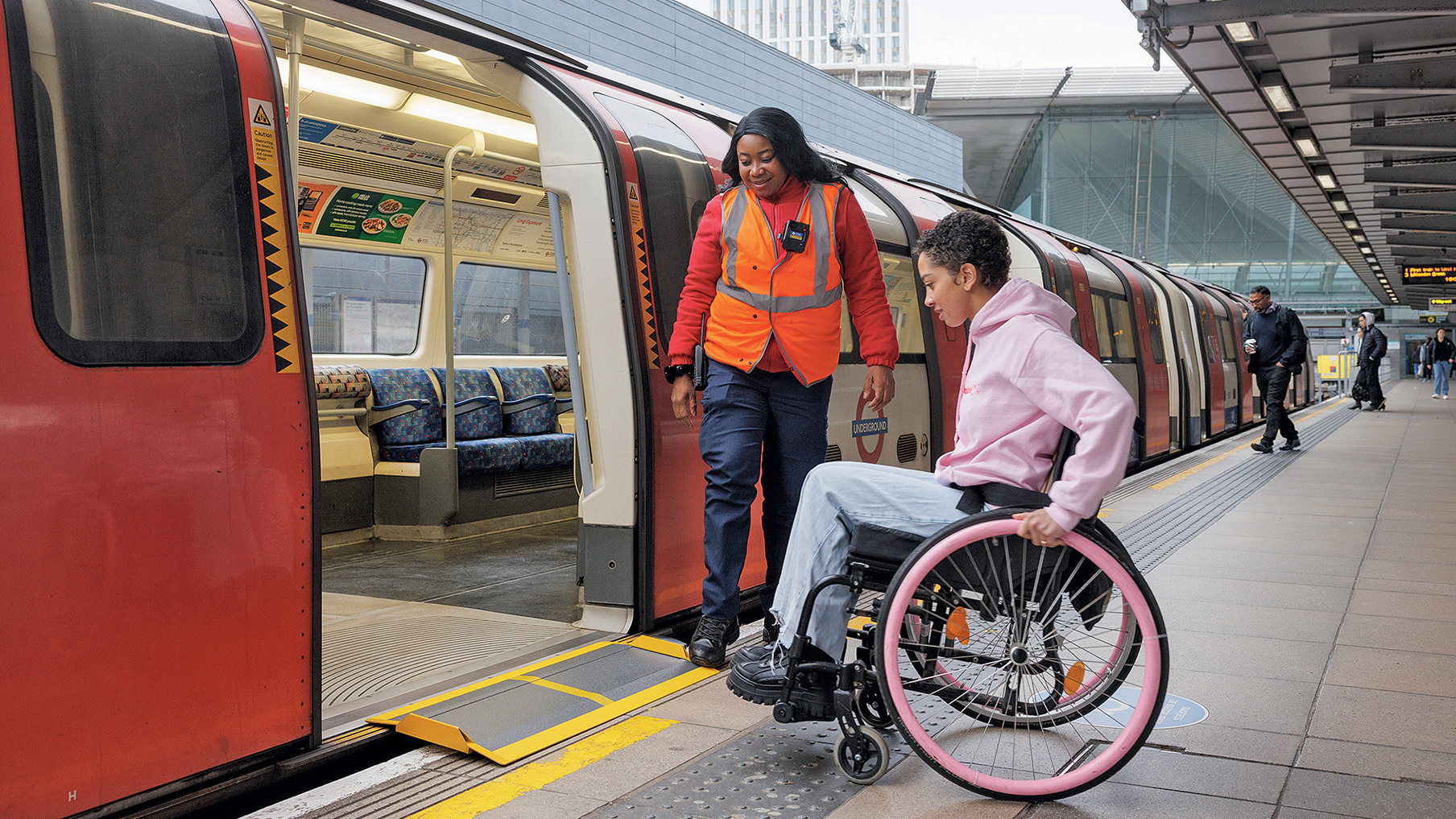 Women in a wheelchair with pink wheels and a pink hoodie going on the tube with some assistance