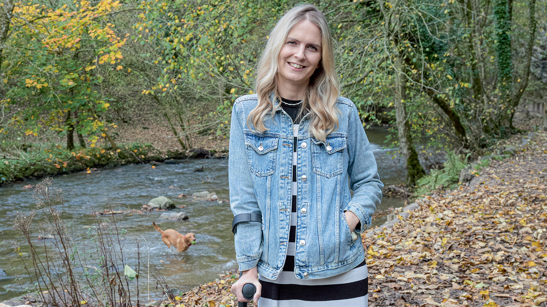 Emma wearing a denim jacket and a dress and holding a walking stick 