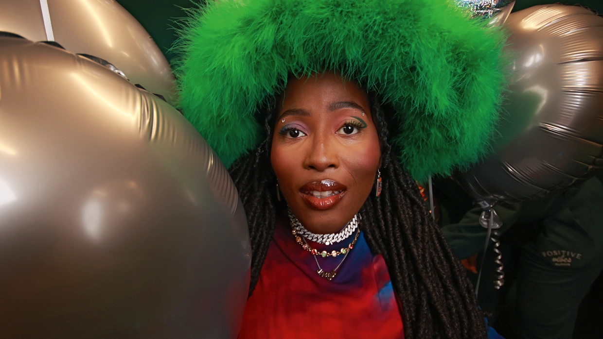 Photo of Miss Jacqui wearing a green fluffy hat, colourful eye shadow and layered necklaces surrounded by balloons 